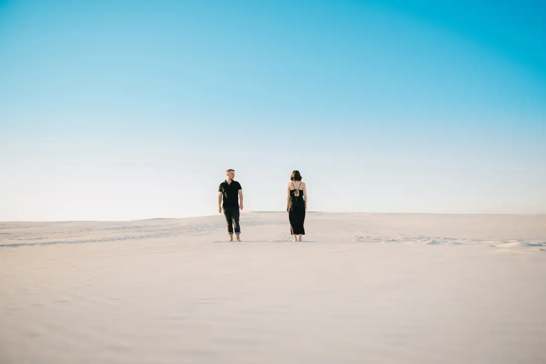 Two people standing apart on pale sand beneath a blue sky
