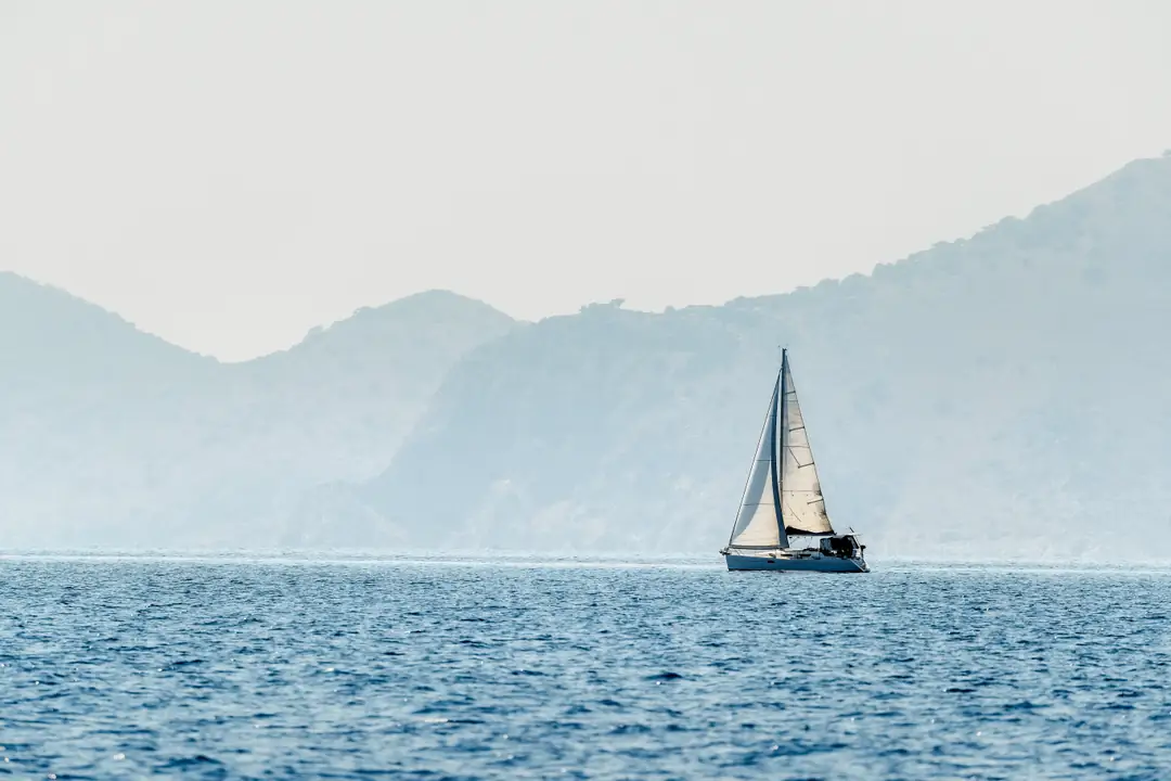 A sailboat on blue water beneath hazy mountains