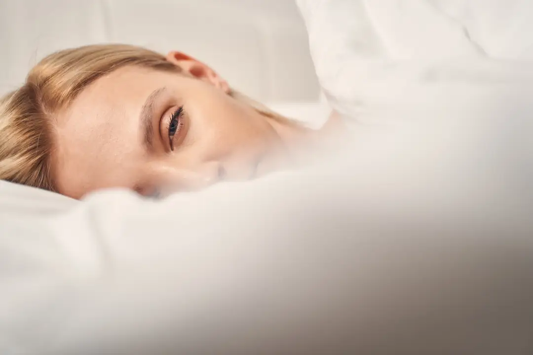 A woman lying awake beneath a cream duvet in soft light